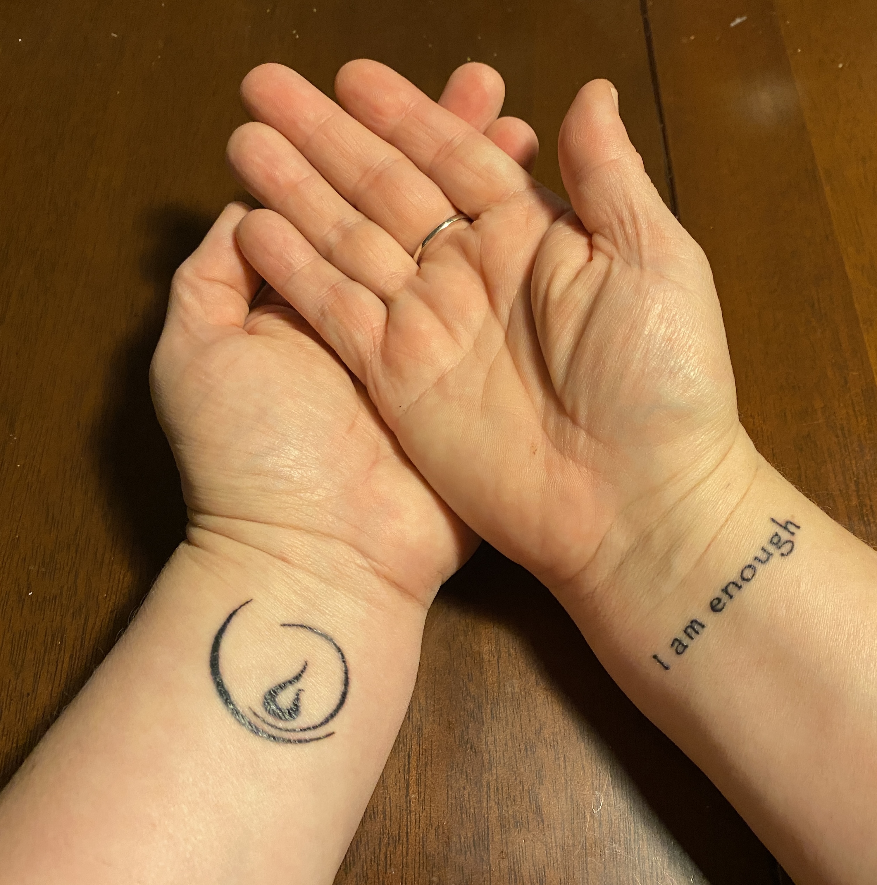 Two hands belonging to Bailey, a white woman, crossed with palms up. One wrist has a chalice flame tattoo, and the other has a tattoo reading I am enough